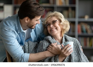 Caring young man covering happy middle aged old mother with plaid, giving cup of hot tea, showing love and devotion at home. Smiling retired woman enjoying peaceful comfort moment with grown son.