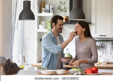 Caring young man in eyewear feeding beloved woman. Loving wife tasting salad, prepared by husband in kitchen, affectionate family couple enjoying cooking together at weekend, preparing breakfast.