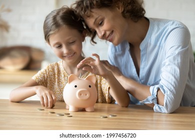 Caring Young Mother Teaching Small Preschool Kid Daughter Saving Money Or Planning Future Purchases, Putting Coins In Small Piggybank In Modern Kitchen, Financial Education For Children Concept.