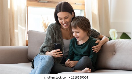 Caring Young Mum Sit On Couch In Living Room With Small Son Watch Funny Cartoon On Smartphone, Happy Mother Or Nanny Relax On Sofa At Home With Little Boy Child Use Cell Browse Internet Together