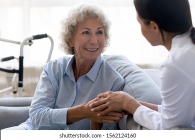 Caring Young Nurse Doctor Carer Helping Holding Hands Of Happy Disabled Handicapped Or Injured Old Adult Elder Woman Having Disability Health Problem Sit At Home With Walking Frame Walker In Hospital
