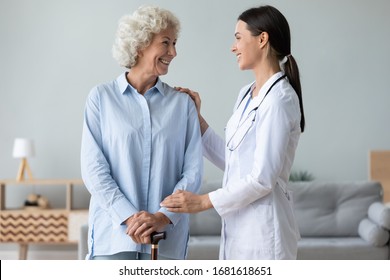 Caring Young Nurse Support Visit Positive Senior Grandmother With Walking Stick At Home, Attentive Female Caregiver Or Doctor Give Help Assist Old Lady Patient In Retirement House Or Clinic