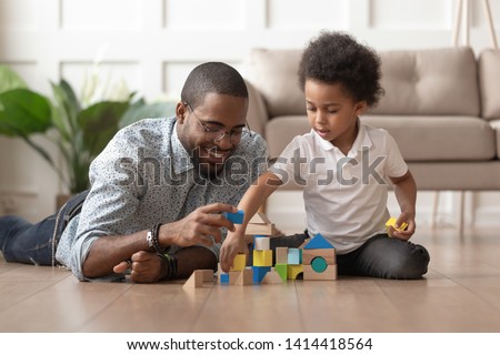 Caring young single black father help cute kid son play on warm floor together, happy african family dad and little child boy having fun building constructor tower from colorful wooden blocks