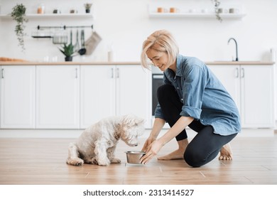 Caring young woman in denim shirt and yoga pants putting dog bowl with breakfast for white furry pet on room floor. Affectionate female owner providing best care for little animal in home interior. - Powered by Shutterstock