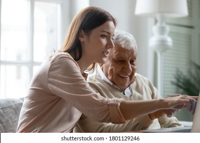 Caring Young Woman Showing Useful Computer Applications To Smiling Old Mature Daddy, Sitting On Sofa At Home. Focused Grown Daughter Teaching Interested Retired Father Using Social Networks On Laptop.