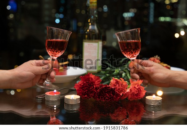 carnation\
flowers and candles in table setting. Hands holding wine and\
blurred colorful city night in\
background