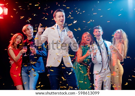 Carnival and party. Emcee with microphone. Group of cheerful friends toasting with wineglasses among confetti.