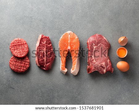 Carnivore or keto diet concept. Raw ingredients for zero carb or low carb diet - burger patties, ribeye, salmon steak, pork, egg on gray stone background.Top view or flat lay.Copy space top and bottom