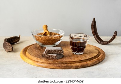 Carob molasses, flour, chocolate and carob pods on wooden cutting board with light grey background and copy space 