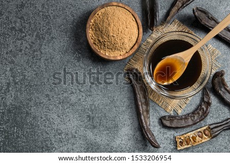 carob molasses in glass bowl and carob powder or flour with carob pods on rustic background, locust bean healthy food, Ceratonia siliqua ( harnup )