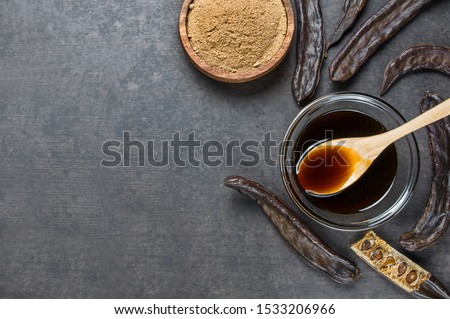 carob molasses in glass bowl and carob powder or flour with carob pods on rustic background, locust bean healthy food, Ceratonia siliqua ( harnup )