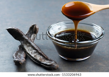 carob molasses in glass bowl and in wooden spoon and carob pods on rustic background, locust bean healthy food, Ceratonia siliqua ( harnup )