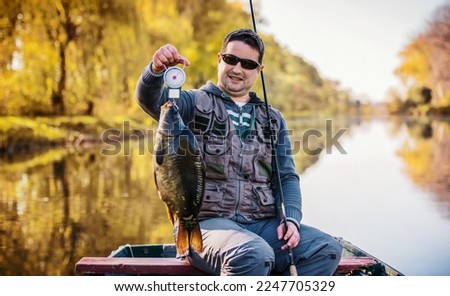 Carp fishing. Fisherman sitting in the boat and measuring carp with fishing scale. Sport and recreation concept