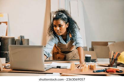 Carpenter america black woman curly hair sketch making notes in work paper while standing at wooden table with laptop computer, young female working learning online at woodshop, Happy Carpenters Day