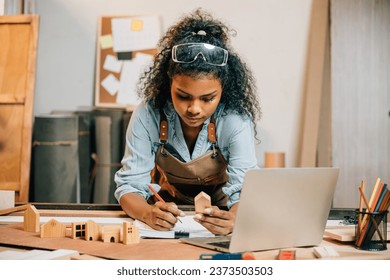Carpenter america black woman curly hair sketch making notes in work paper while standing at wooden table with laptop computer, young female working learning online at woodshop, Happy Carpenters Day