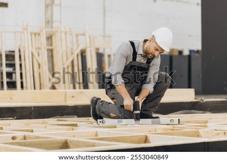 Carpenter assembling prefabricated house module using spirit level and screwdriver, working on wooden frame in bright carpentry workshop