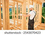 Carpenter building wooden frame house. Man measures the distance with tape measure while dressed in workwear and a helmet.