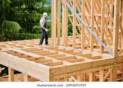 Carpenter constructing house with wooden frame. Man measures distance using tape measure, wearing protective clothing and helmet. Concept of environmentally sound, modern construction. - Powered by Shutterstock