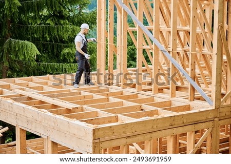 Carpenter constructing house with wooden frame. Man measures distance using tape measure, wearing protective clothing and helmet. Concept of environmentally sound, modern construction.