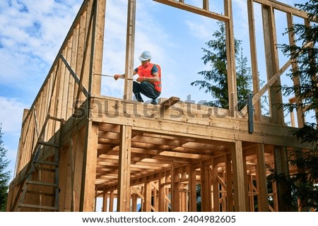 Carpenter constructing timber-framed two-story building. Man measures distances with measuring tape while dressed in workwear and hard hat. Ideology of contemporary, eco-friendly building practices.