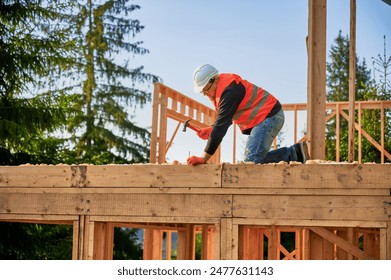 Carpenter constructing two-story wooden frame house near forest. Bearded man hammering nails into the structure, wearing protective helmet, construction vest. Concept of modern ecological construction - Powered by Shutterstock