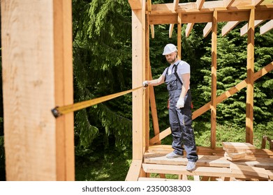Carpenter constructing wooden skeleton building. Man measures distances with tape measure while wearing work attire and helmet. The concept of modern, environmentally-friendly construction. - Powered by Shutterstock