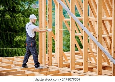 Carpenter constructing wooden-framed dwelling. Man measures distances with measuring tape while wearing work attire and headgear. The idea behind contemporary, eco-friendly building practices. - Powered by Shutterstock