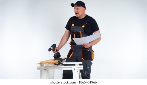 Carpenter with a cordless screwdriver in his hands. A man in work clothes holds drawings in his hands and communicates with a colleague backstage. Joiner in a gray jumpsuit and a baseball cap. - Powered by Shutterstock