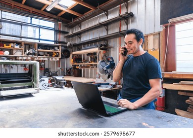 Carpenter and craftsman managing orders using laptop and talking on phone in workshop