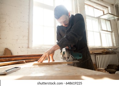 Carpenter Cutting Wooden Board On Workbench With Handle Circular Saw. Entrepreneur Working On His Small Woodworking Venture. Woodworker Works On Local Lumber Production Or Custom Furniture Manufacture