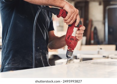 Carpenter is drilling a hole in a white wood plank using a power drill in a workshop. The carpenter is wearing a black t shirt and is using a red corded drill - Powered by Shutterstock