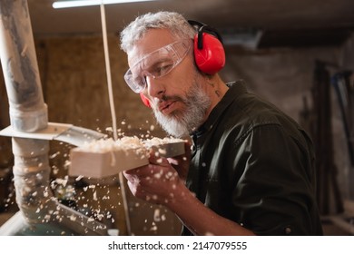 carpenter in goggles and protective earmuffs blowing sawdust out of plank - Powered by Shutterstock