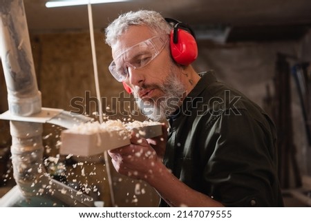 carpenter in goggles and protective earmuffs blowing sawdust out of plank