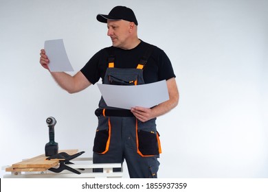 Carpenter in a gray jumpsuit and a baseball cap. Joiner examines the drawings. A man stands in a carpenter shop. The concept of woodworking. Manufacturing of furniture to order. - Powered by Shutterstock