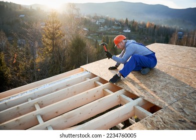 Carpenter hammering nail into OSB panel on the roof top of future cottage at sunrise. Man worker building wooden frame house. Carpentry and construction concept. - Shutterstock ID 2234760005