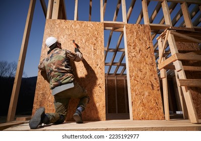 Carpenter hammering nail into OSB panel on the wall of future cottage. Man worker building wooden frame house. Carpentry and construction concept. - Shutterstock ID 2235192977
