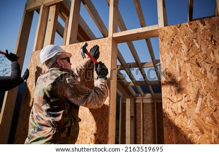 Carpenter hammering nail into OSB panel on the wall of future cottage. Man worker building wooden frame house in the Scandinavian style barnhouse. Carpentry and construction concept.