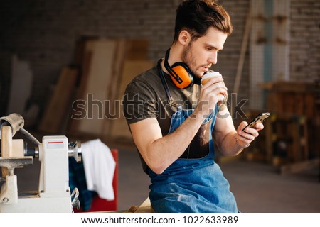 Carpenter Man using smart phone and holding cup of coffee in front of table with tools at workshop