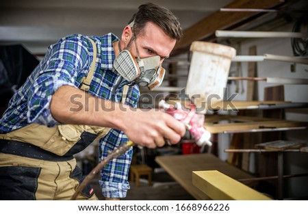 Carpenter with mask using paint spray gun to painting wooden plank in carpentry workshop