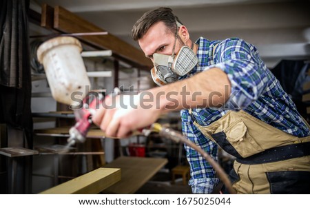 Carpenter with mask using paint spray gun to painting wooden plank in carpentry workshop
