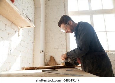 Carpenter In Protective Glasses Polishes Wooden Board With Handle Belt Sanding Machine On Workbench In Workshop. Small Business Owner Working In His Manufacturing. Joiner Made Custom Wooden Furniture