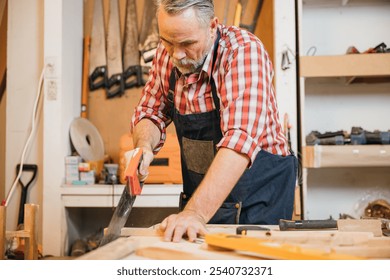 A carpenter in a red plaid shirt and blue overalls saws a piece of wood with a handsaw on a workbench, concentrating on precise cuts in the woodworking project, National Carpenters Day - Powered by Shutterstock