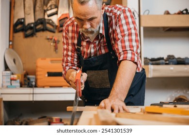 A carpenter in a red plaid shirt and blue overalls saws a piece of wood with a handsaw on a workbench, concentrating on precise cuts in the woodworking project, National Carpenters Day - Powered by Shutterstock