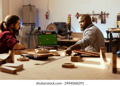 Carpenter talking with client about commissioned furniture assembling job, presenting on mockup laptop. Cabinetmaker shows customer blueprint on CAD software on green screen notebook - Powered by Shutterstock