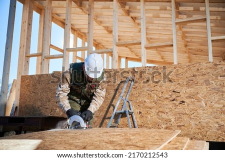 Carpenter using circular saw for cutting wooden OSB board. Man worker building wooden frame house on pile foundation. Carpentry concept.