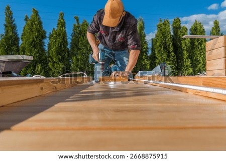 A carpenter is using a drill to secure wooden planks while constructing a deck outdoors on a sunny day. Lush trees are visible in the background.