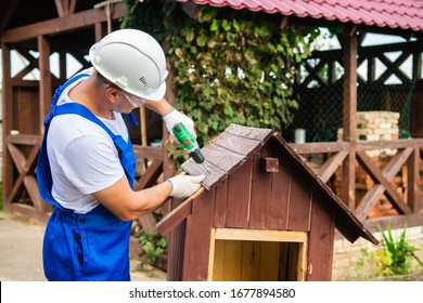 Carpenter Using Electric Screwdriver. Man Builds A Roof Of Wooden Planks Of A Small House. Building Dog Booth. Countryside House.