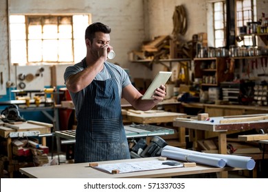 carpenter using his tablet and drinking a coffee in a dusty workshop - Powered by Shutterstock