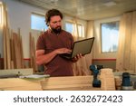 Carpenter is using laptop in his workshop, managing orders and checking inventory, surrounded by wood planks and tools, ensuring efficient workflow and client satisfaction