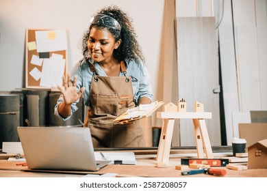 Carpenter woman in a workshop holding furniture design sketches, presenting her project to a customer using a laptop, surrounded by unfinished wooden pieces and tools, craftsmanship and creativity